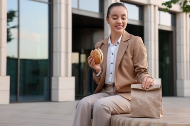 Photo of Happy businesswoman with hamburger having lunch on bench outdoors