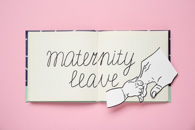 Photo of Notebook with phrase Maternity Leave and paper cutout of hands on pink background, top view