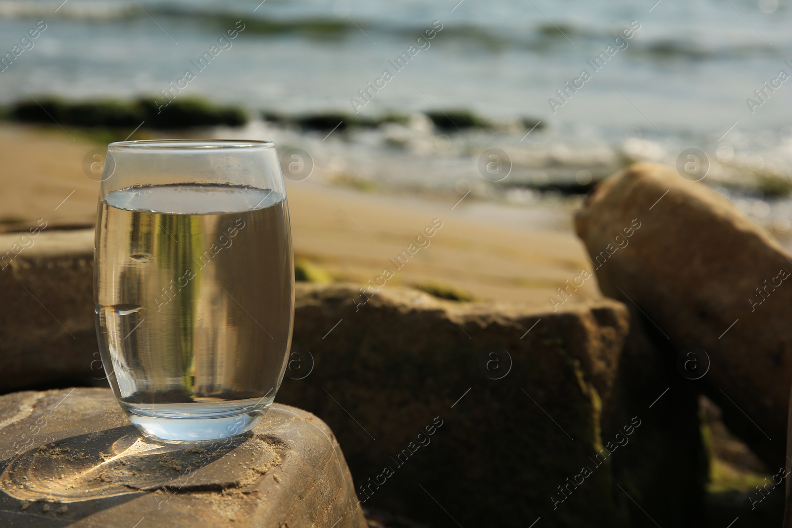 Photo of Glass of fresh water on stone near sea, space for text