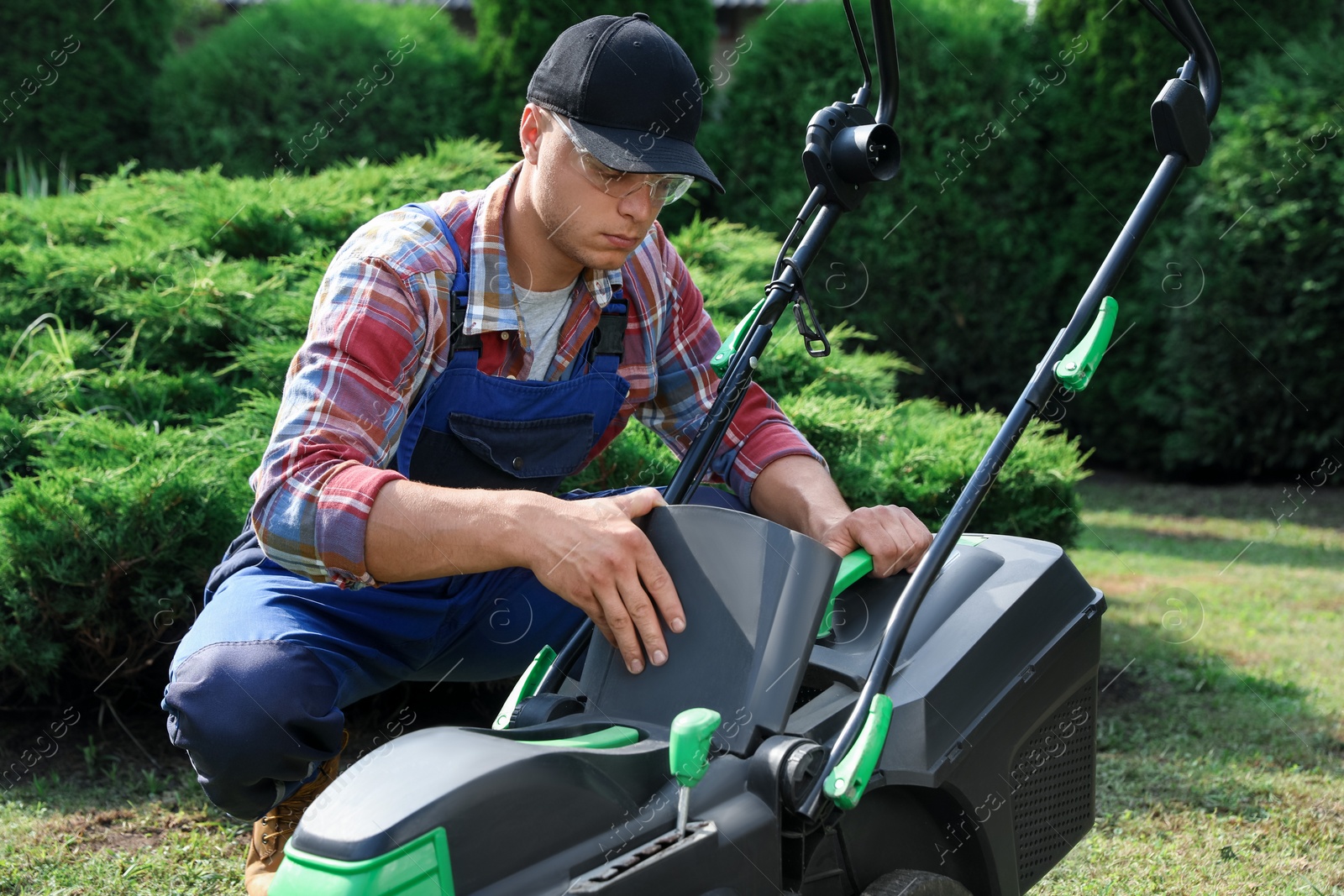 Photo of Cleaning lawn mower. Young man detaching grass catcher from device in garden