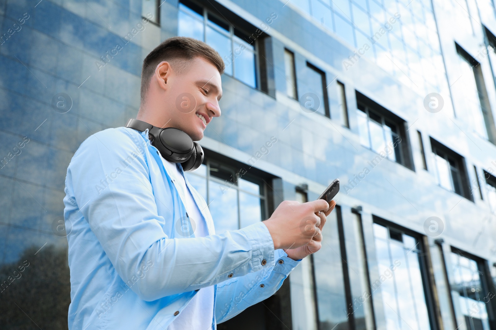 Photo of Smiling man with headphones using smartphone near building outdoors. Space for text