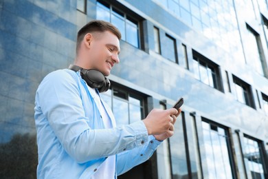 Photo of Smiling man with headphones using smartphone near building outdoors. Space for text