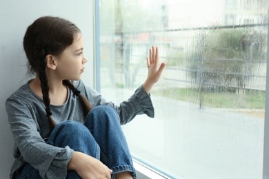 Photo of Sad little girl near window indoors on rainy day