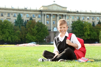 Photo of Cute boy with school stationery sitting on green lawn outdoors