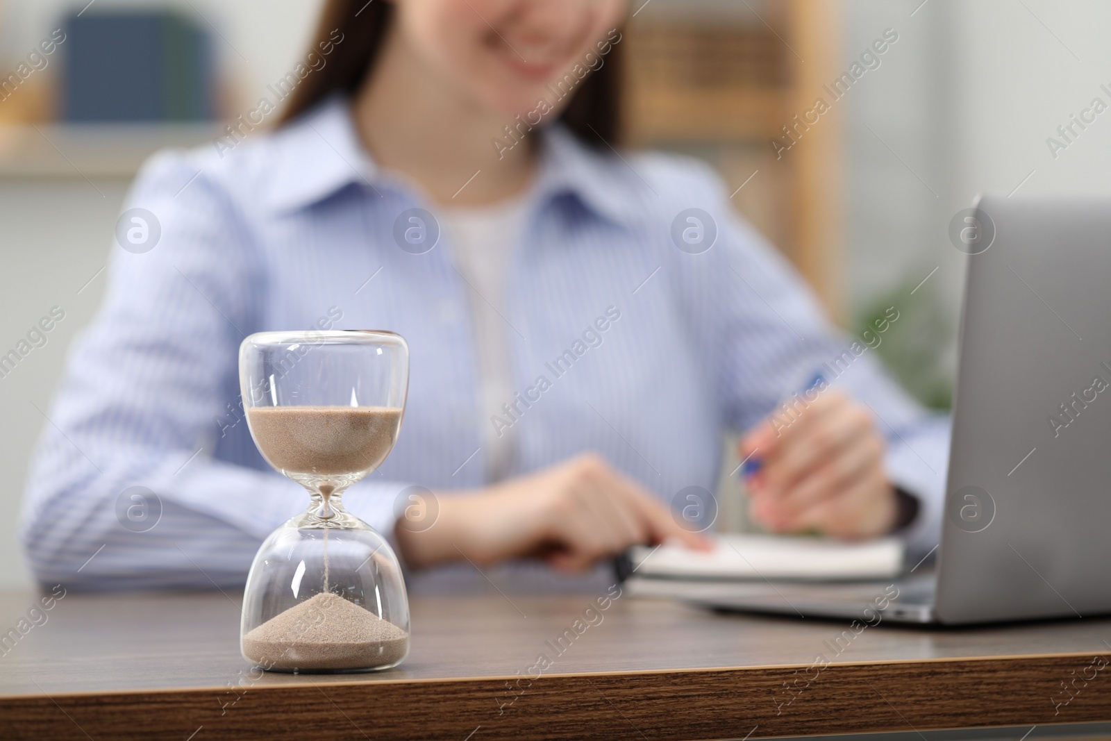 Photo of Hourglass with flowing sand on desk. Woman taking notes while using laptop indoors, selective focus