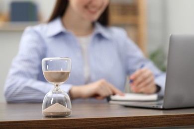 Hourglass with flowing sand on desk. Woman taking notes while using laptop indoors, selective focus