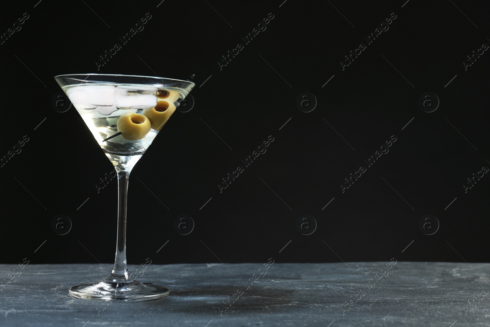 Photo of Martini cocktail with ice and olives on grey table against dark background. Space for text