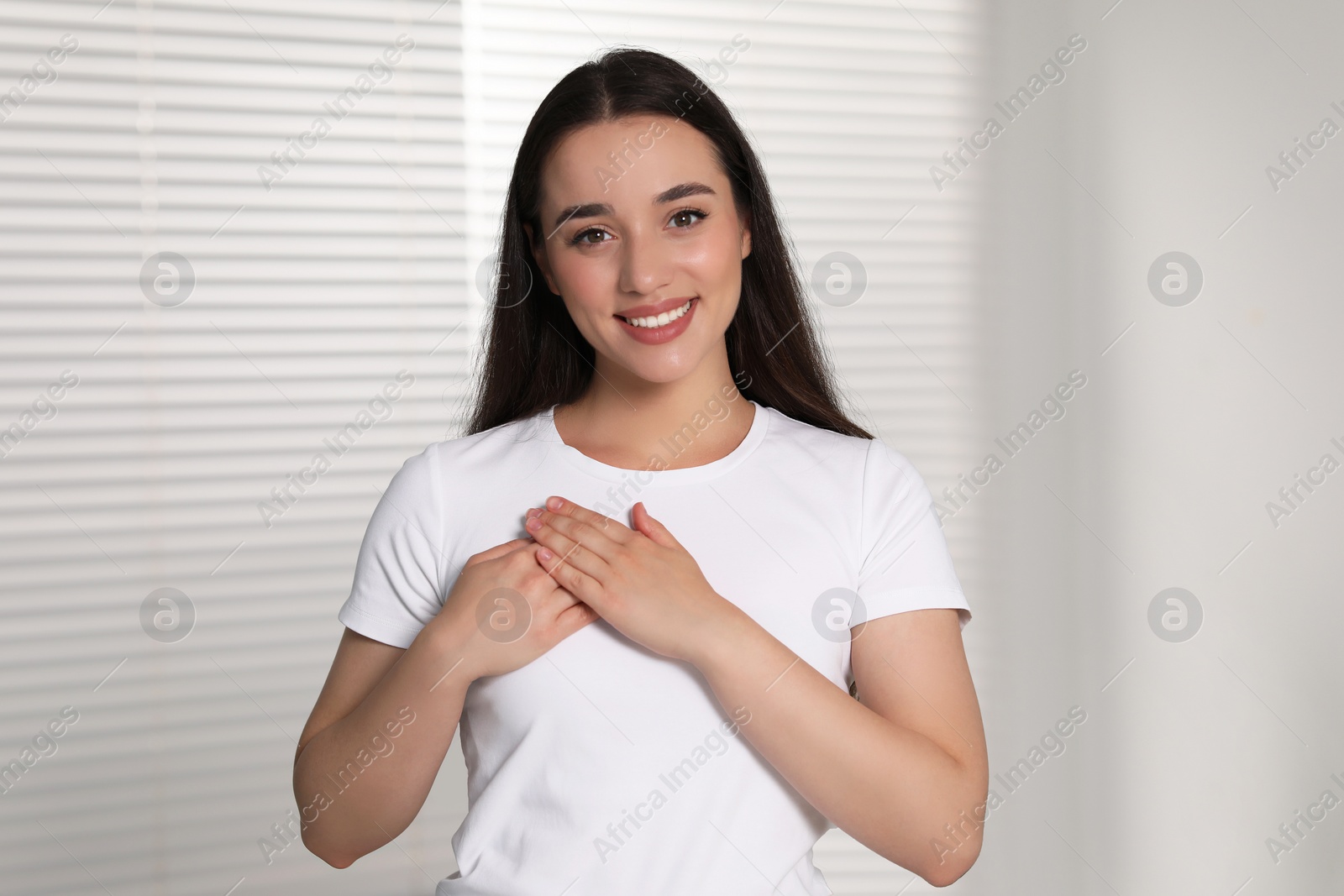 Photo of Thank you gesture. Beautiful grateful woman with hands on chest indoors