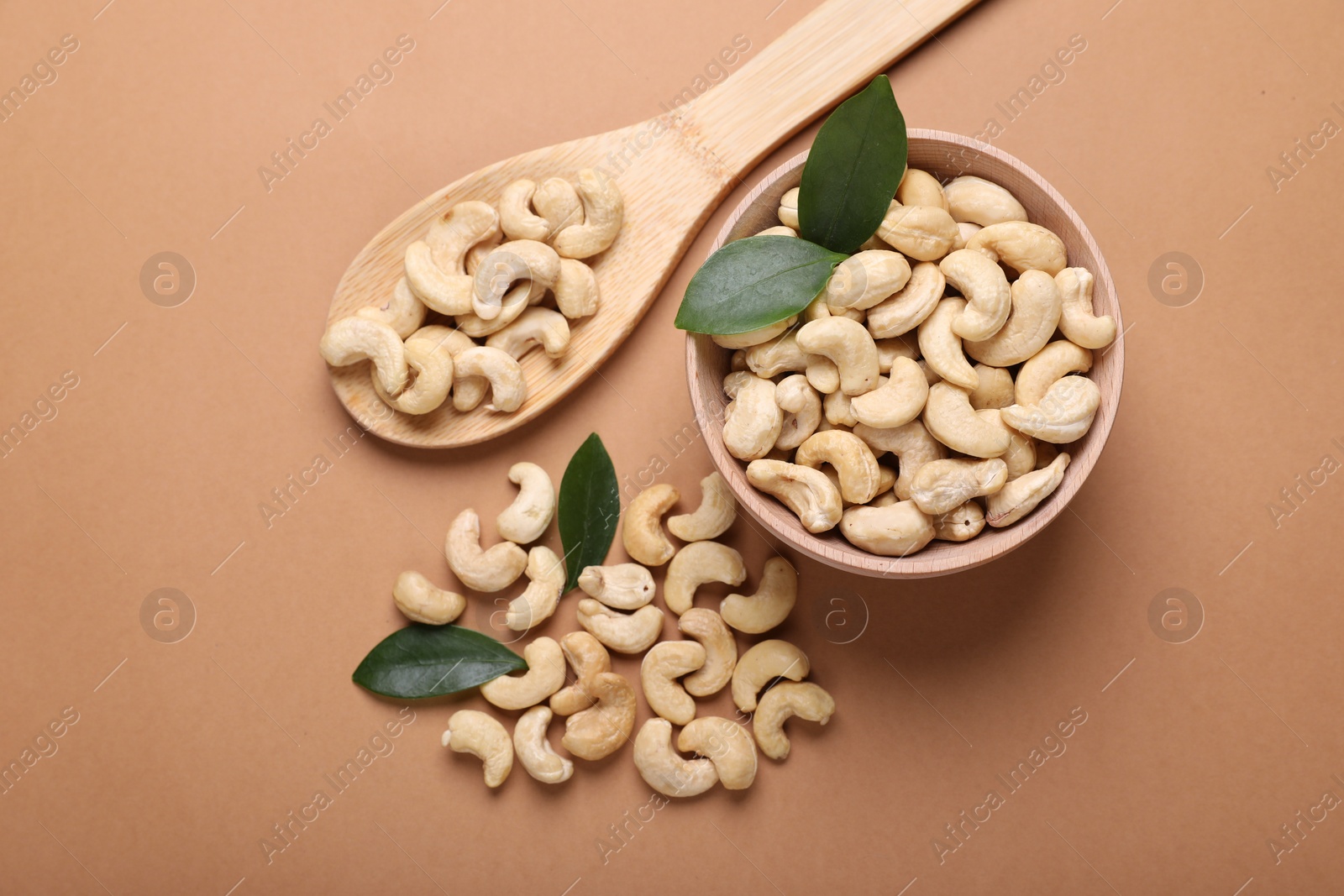 Photo of Tasty cashew nuts and green leaves on pale brown background, flat lay