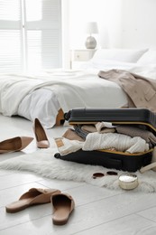 Open suitcase full of clothes, shoes and fashionable accessories on floor in bedroom