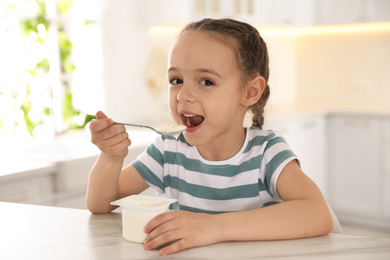 Cute little girl eating tasty yogurt at table in kitchen