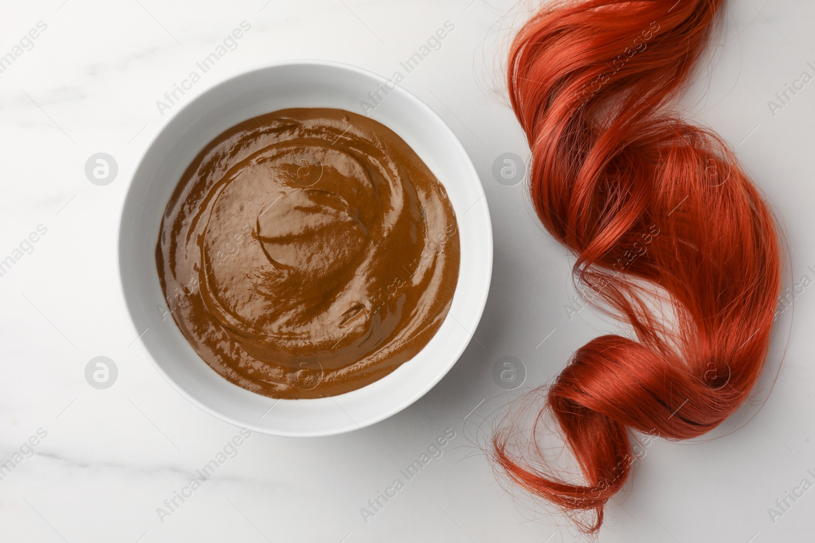 Photo of Bowl of henna cream and red strand on white marble table, flat lay. Natural hair coloring
