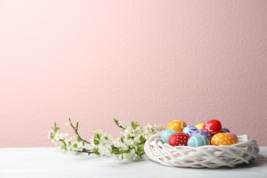 Photo of Painted Easter eggs in nest and blossoming branches on table against color background. Space for text