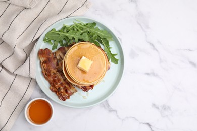 Tasty pancakes with butter, fried bacon and fresh arugula on white marble table, flat lay. Space for text