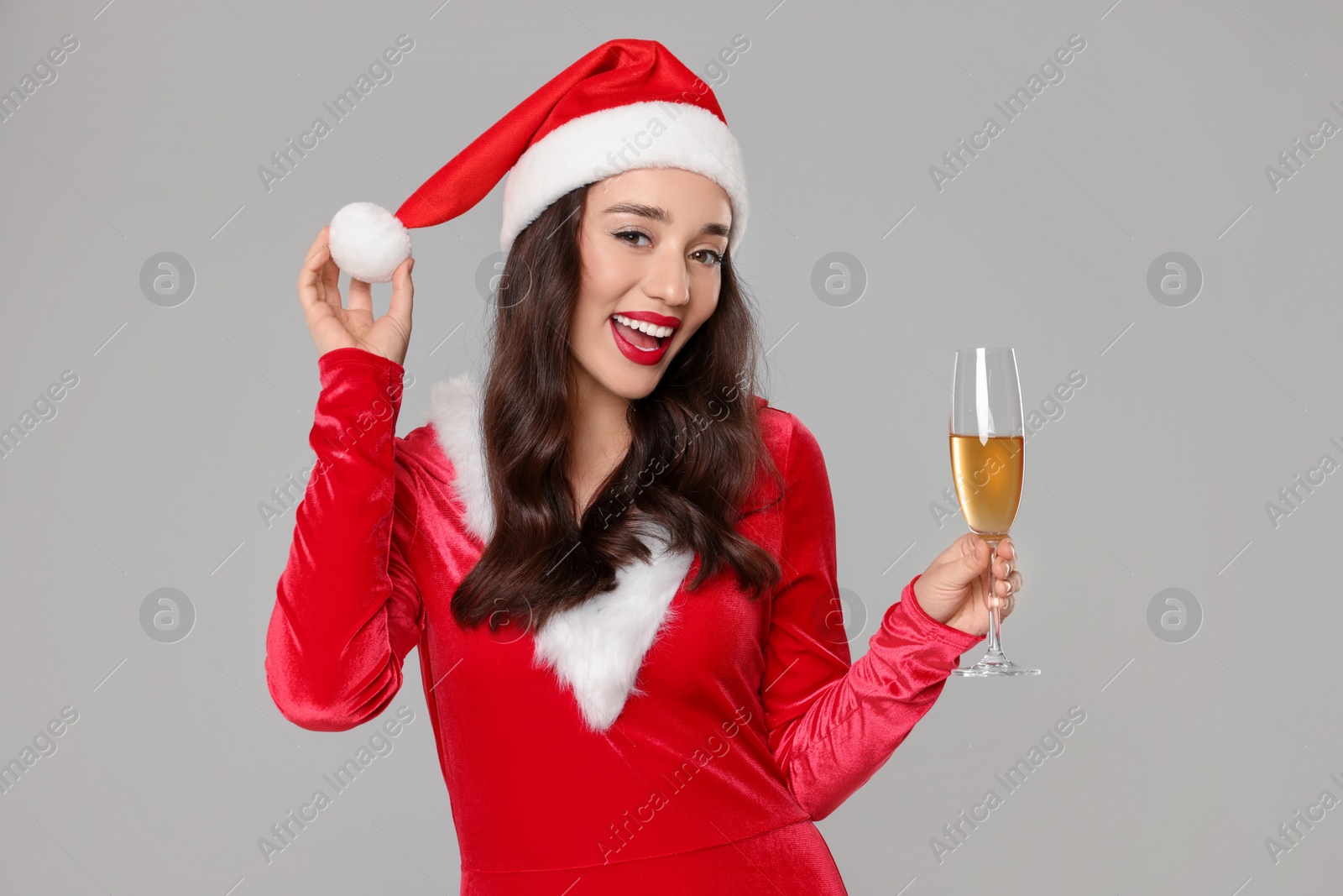 Photo of Christmas celebration. Beautiful young woman in red dress and Santa hat with glass of champagne on grey background