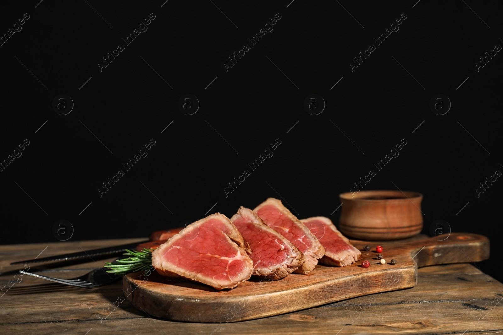 Photo of Board with roasted meat and rosemary on wooden table against black background. Space for text