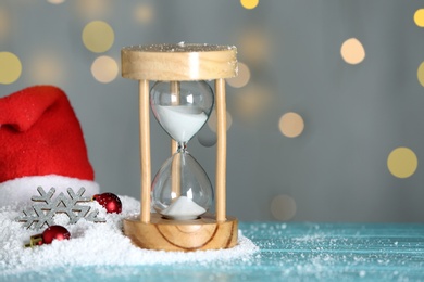 Hourglass with snow and decor on light blue wooden table against blurred lights, space for text. Christmas countdown