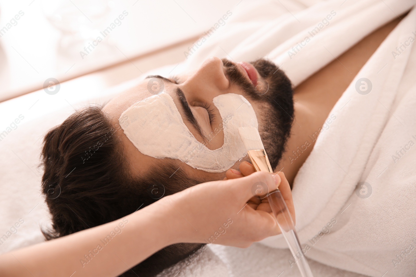 Photo of Cosmetologist applying mask on man's face in spa salon