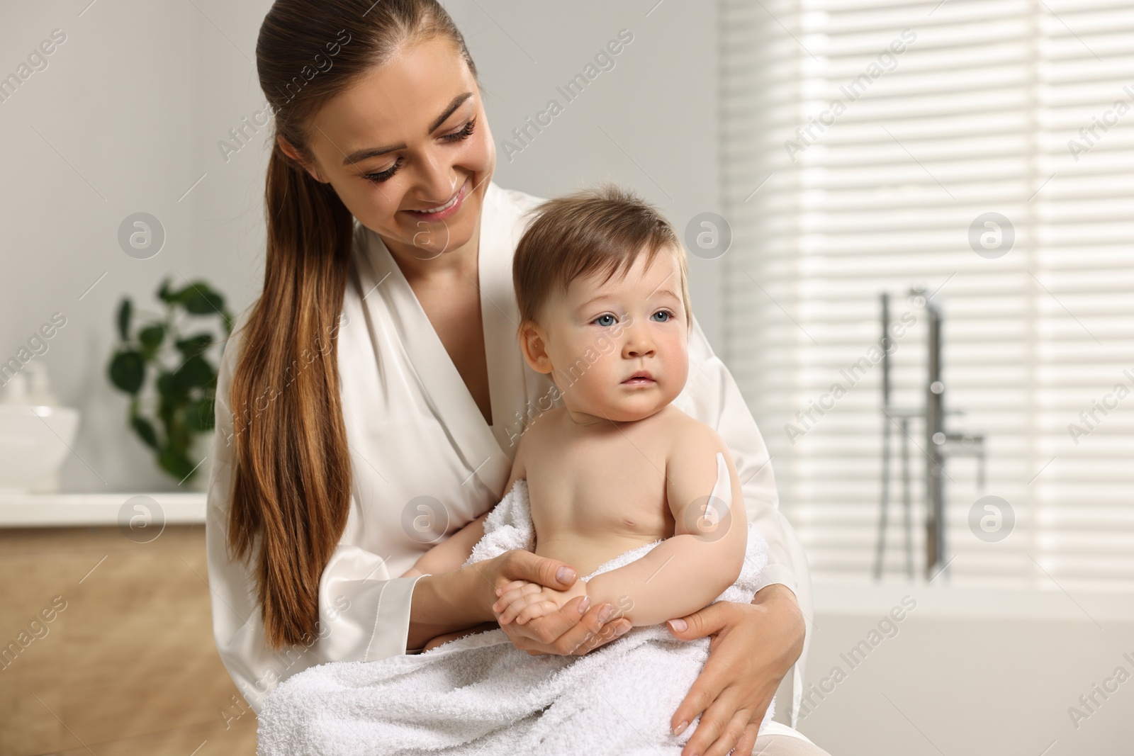 Photo of Happy mother holding her baby with body cream on skin in bathroom