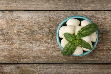 Photo of Tasty mozzarella balls and basil leaves in bowl on wooden table, top view. Space for text