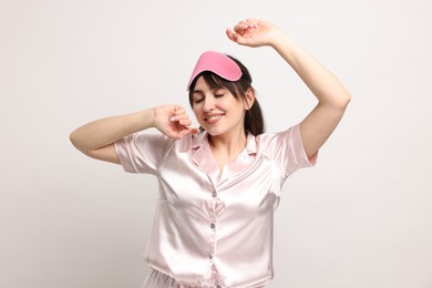 Happy woman in pyjama and sleep mask on light grey background