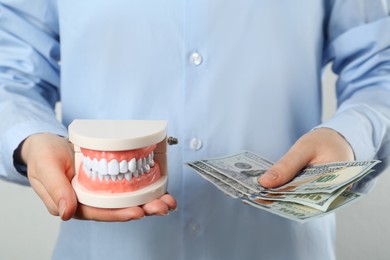 Woman holding educational dental typodont model and dollar banknotes on light background, closeup. Expensive treatment