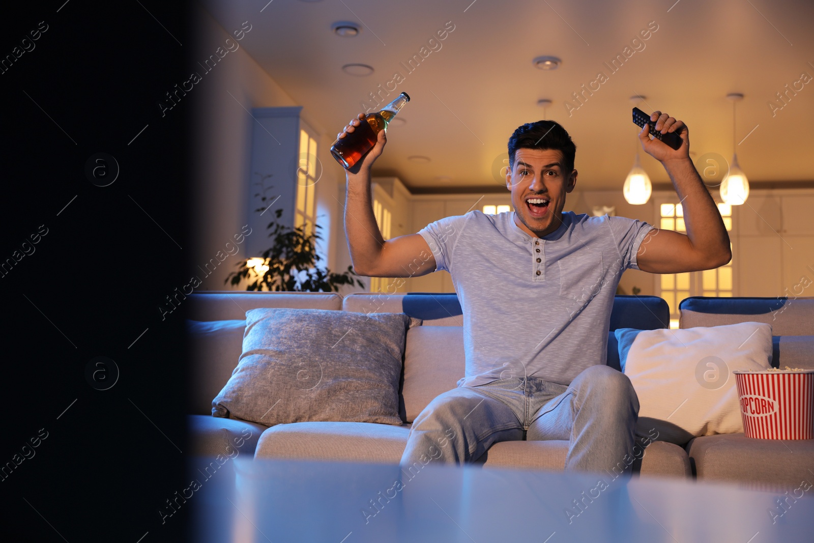 Photo of Man watching movie with popcorn and bottle of beer on sofa at night