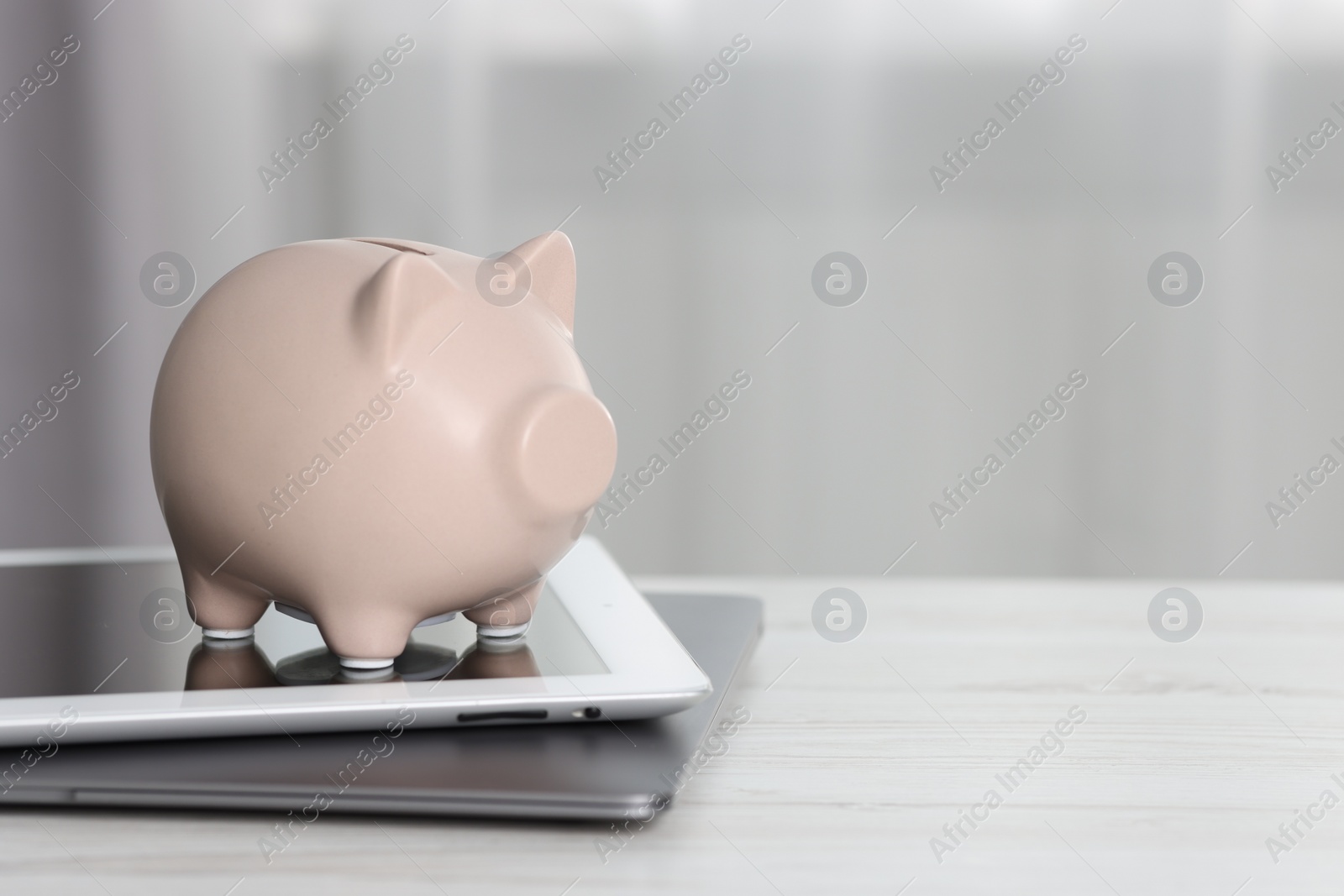 Piggy bank, tablet and laptop on white table indoors. Space for text Photo of Piggy bank, tablet and laptop on white table indoors. Space for text