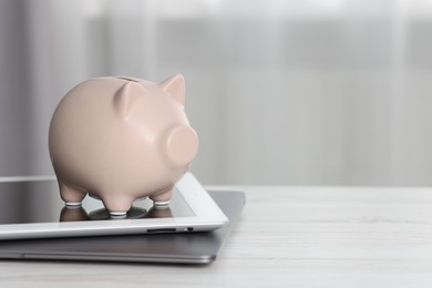 Photo of Piggy bank, tablet and laptop on white table indoors. Space for text