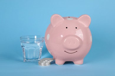 Photo of Water scarcity concept. Piggy bank, coins and glass of drink on light blue background