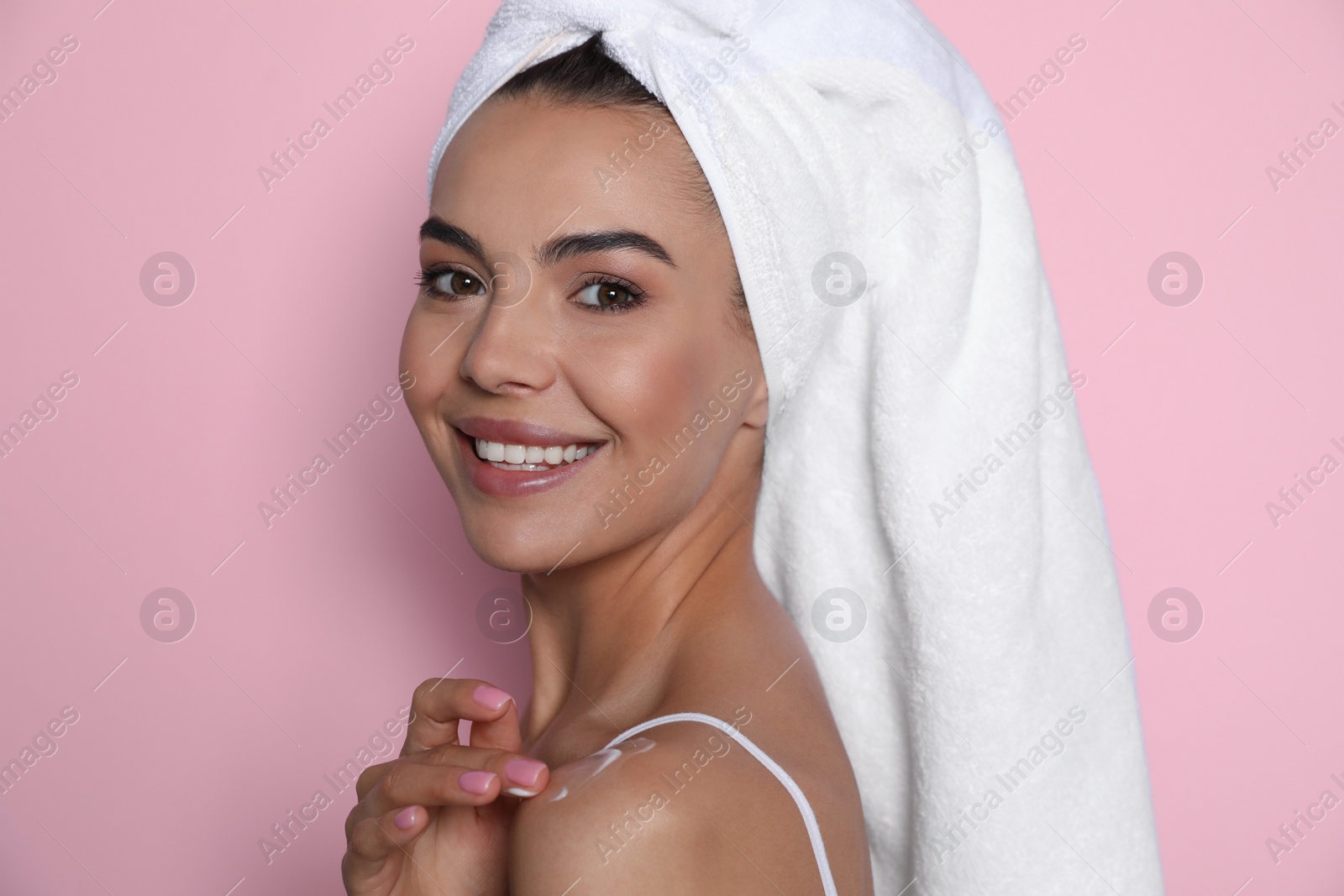 Photo of Beautiful young woman with towel applying cream on shoulder against pink background