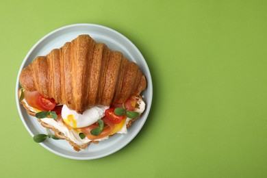 Tasty croissant with fried egg, tomato and microgreens on green background, top view. Space for text