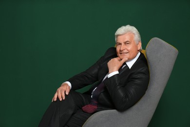 Happy mature businessman sitting in armchair against green background, space for text
