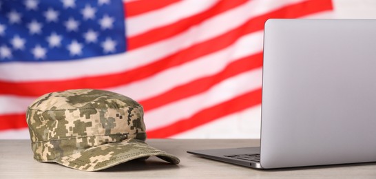 Image of Military education. Cap and laptop on wooden table against flag of USA