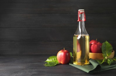 Bottle of delicious cider and apples with green leaves on black wooden table, space for text