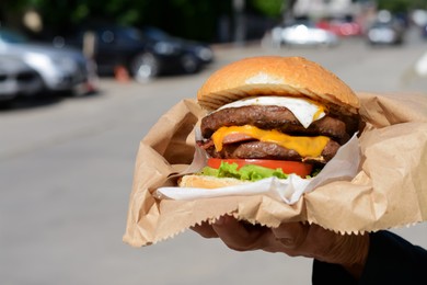 Photo of Woman holding delicious burger in paper wrap on city street, closeup. Space for text