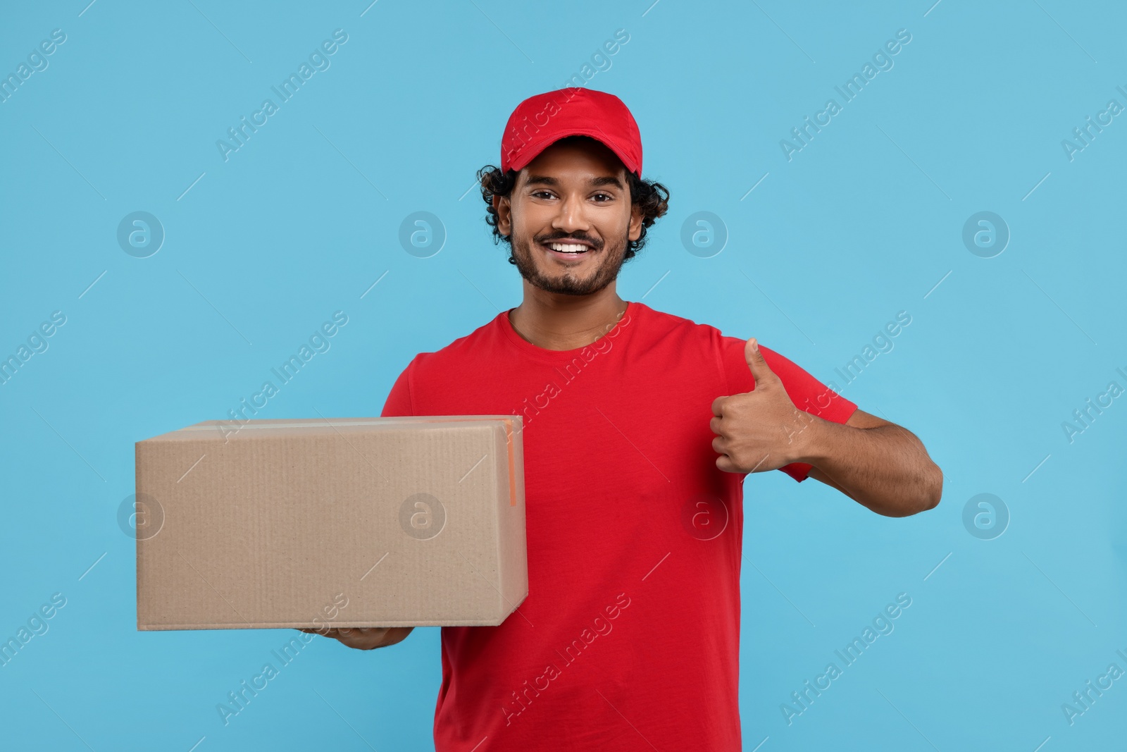 Photo of Happy young courier with parcel showing thumb up on light blue background