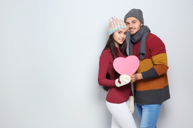 Photo of Young happy couple in warm clothes holding pink heart on light background. Christmas celebration