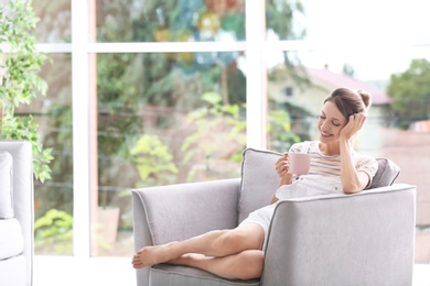Photo of Young woman with cup of aromatic coffee sitting in armchair near window at home