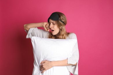 Photo of Young tired woman with sleeping mask and pillow yawning on pink background