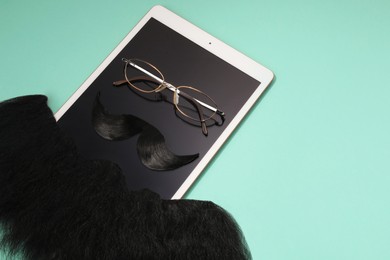 Photo of Artificial moustache, beard, tablet and glasses on turquoise background, above view. Space for text
