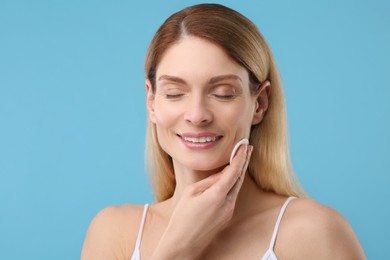 Beautiful woman removing makeup with cotton pad on light blue background