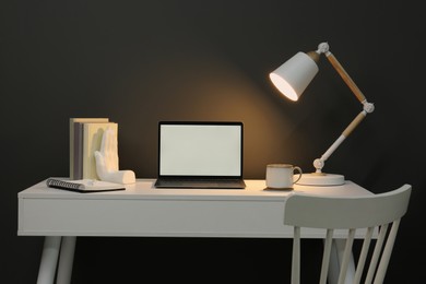 Stylish workplace with laptop, lamp, cup and decor on white table near grey wall