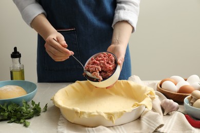 Woman putting meat into baking dish with dough to make pie at light grey table, closeup