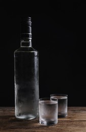 Photo of Bottle of vodka and shot glasses on wooden table against black background. Space for text