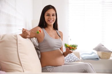 Photo of Young pregnant woman with bowl of vegetable salad in living room. Taking care of baby health