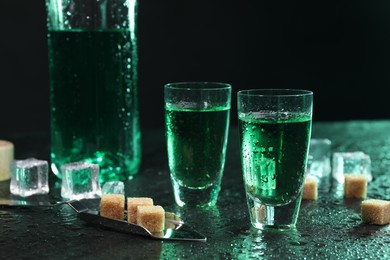 Photo of Absinthe in shot glasses, spoon, brown sugar and ice cubes on gray table against dark background, closeup. Alcoholic drink