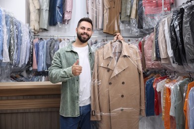 Photo of Dry-cleaning service. Happy man holding hanger with coat in plastic bag and showing thumb up indoors
