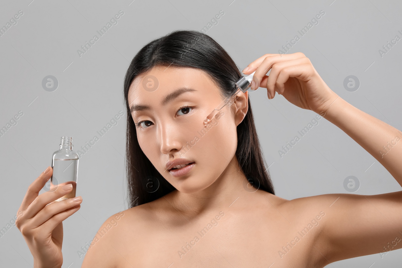 Photo of Beautiful young woman applying cosmetic serum onto her face on grey background