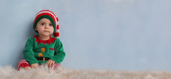 Image of Cute baby wearing elf costume on fluffy carpet near light blue wall, banner design with space for text. Christmas celebration